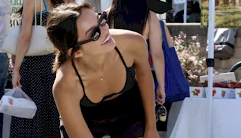 A woman wearing sunglasses and a black tank top smiles while leaning forward at an outdoor market stand, with people and bags visible in the background.