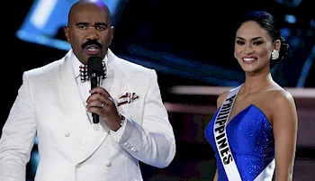 A man in a white suit holds a microphone while standing next to a smiling woman wearing a blue gown and a sash that reads Philippines at a pageant event.