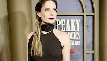 A woman in a black sleeveless dress with a scarf around her neck stands in front of a Peaky Blinders promotional backdrop. She has long, light brown hair styled in a loose braid.