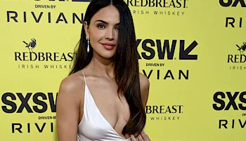 A dark haired woman smiles as she looks into the camera. She is wearing a light silk dress and is posing against a yellow background with various brand names written in black.
