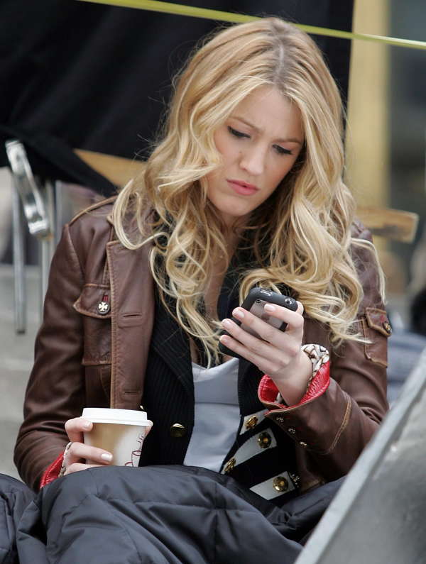 A woman with long blonde hair sits outdoors, looking at her smartphone. She is wearing a brown leather jacket and holding a coffee cup in her other hand. She appears focused on the phone screen.