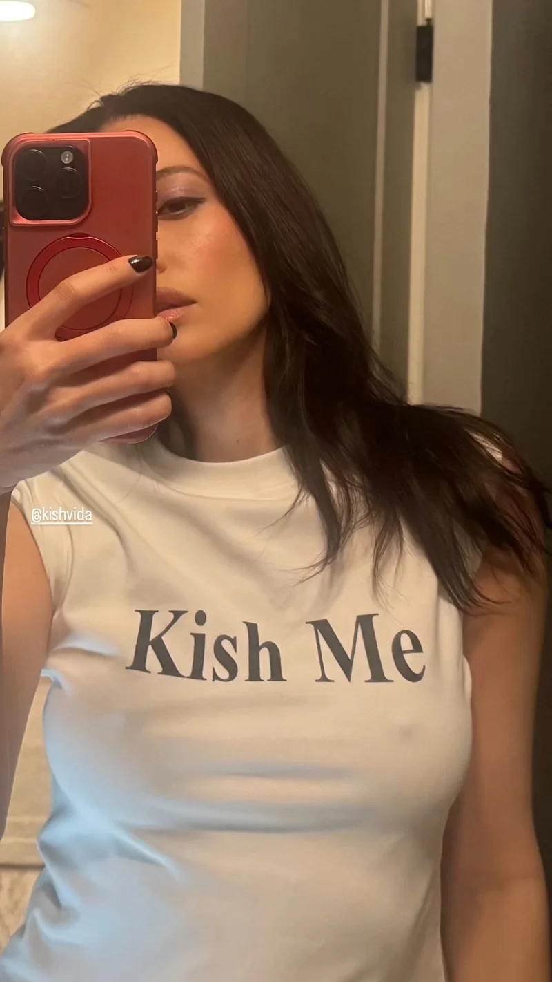 ChristianSerratosKishMe A woman with long dark hair takes a mirror selfie with her phone, partially covering her face. She is wearing a white sleeveless top with the text Kish Me printed on it.
