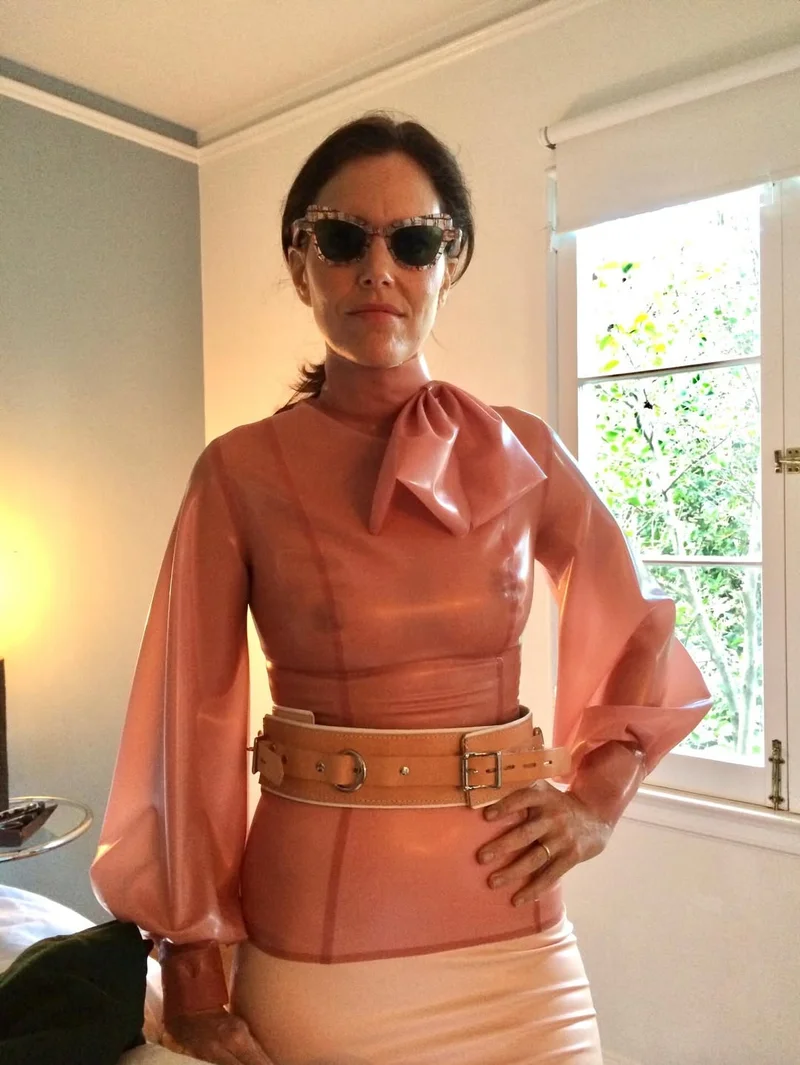 IoneSkyePinkLatexSeeThrough A woman wearing sunglasses and a sheer pink blouse with a large bow and puffy sleeves stands indoors with one hand on her hip, next to a window with greenery visible outside.