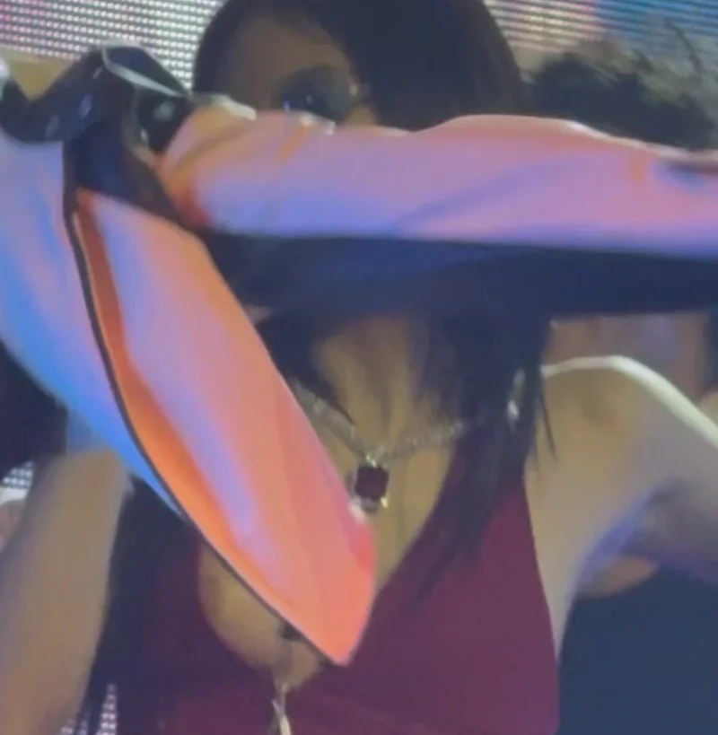 Asian woman wearing glasses and a red top is partially obscured by a white and orange jacket in the foreground, with colorful lighting and a blurred background #3.