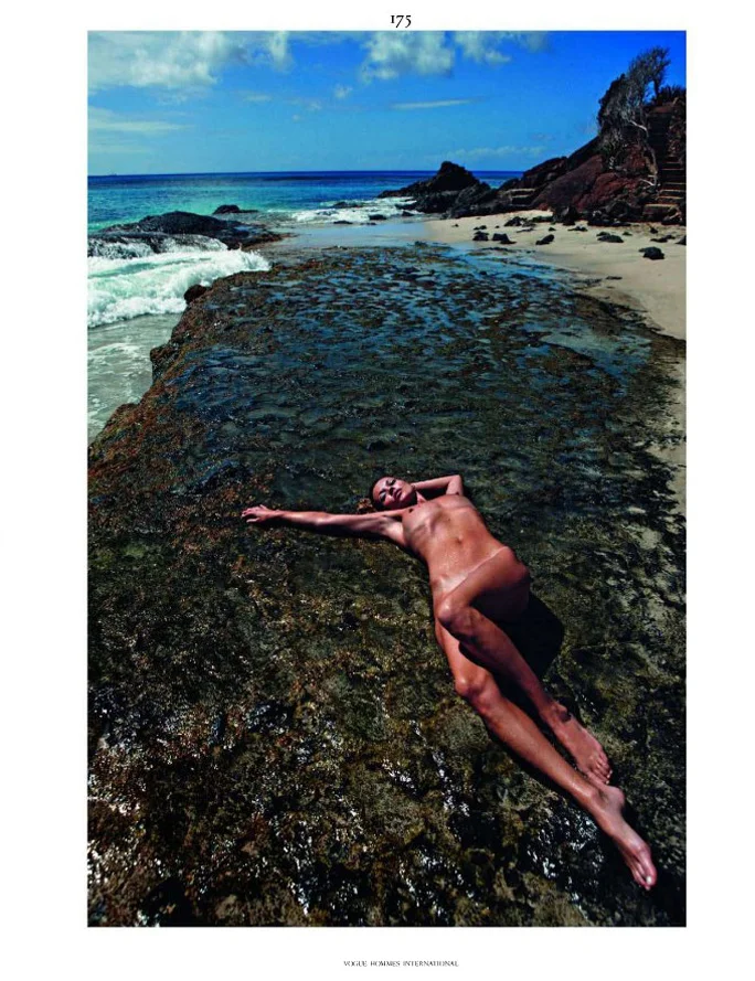 Kate Moss lies nude on a rocky outcrop by the shore, with waves gently approaching from the left and a sandy beach and blue sky in the background.
