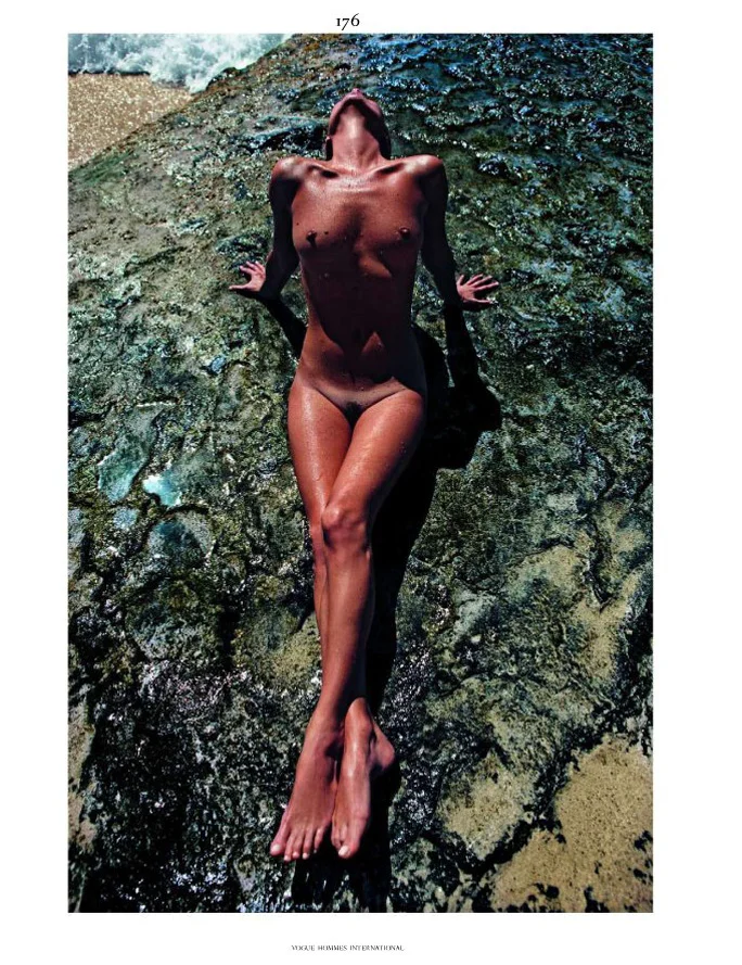 Kate Moss lies nude on a rocky, moss-covered surface by the water, basking in sunlight with arms extended back and legs straight, head tilted upward, and waves visible in the corner.