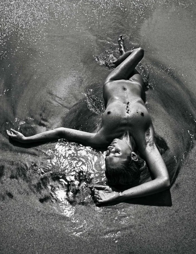 Kate Moss lies nude on wet sand at the edge of the water, arms outstretched and eyes closed, with sunlight glistening on their body and the surrounding sand. The image is in black and white.