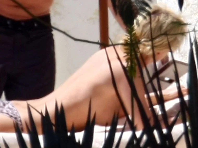 A woman with blonde hair is lying face down on a lounge chair outdoors, possibly sunbathing. They are topless, and leafy plants partially obscure the view. Another person stands nearby.