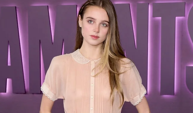 A young woman with long brown hair poses in front of a purple wall with large letters. She is wearing a sheer, light pink blouse with a white collar and has a neutral expression.