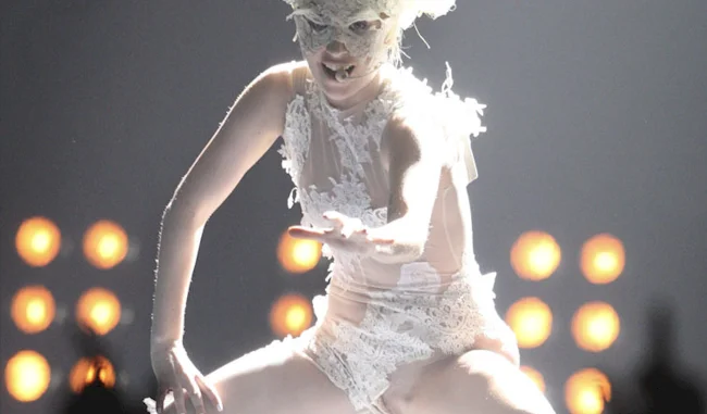 Lady GaGa wearing a mask and semi-sheer costume while singing on a stage with lights in the background