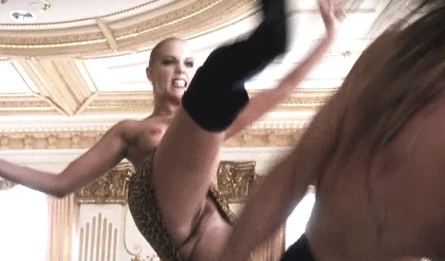Screen capture of a topless Elizabeth Berkely making a fierce face and is in mid-kick. Her vagina is on full display.