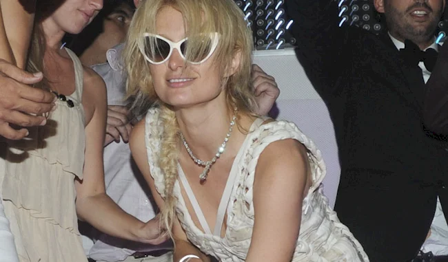 Paris Hilton wearing large white sunglasses, a white sleeveless dress, and a necklace poses with a smile in a crowded, lively indoor party setting. Other people are visible around her.