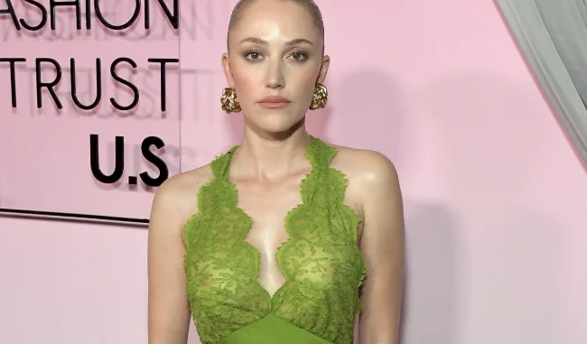 A woman with light skin and blonde hair in a sleek bun poses in front of a pink backdrop, wearing a sleeveless green lace dress and gold earrings. Black text on the wall reads FASHION TRUST U.S.