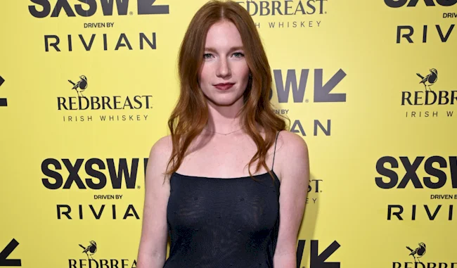 A woman with long red hair in a black dress poses in front of a yellow SXSW event backdrop featuring sponsor logos, including Redbreast whiskey and Rivian.