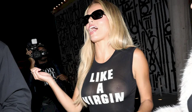 Brooks Nader with long blonde hair and sunglasses walks at night, wearing a sheer black top displaying the text LIKE A VIRGIN. She is mid-gesture, and a photographer is in the background capturing the scene.