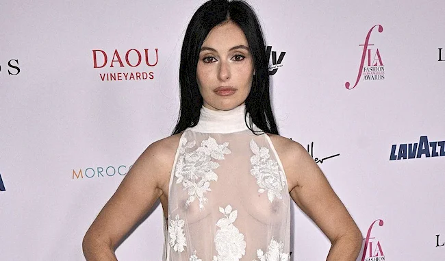 A woman with long dark hair poses on a red carpet, wearing a sheer white dress with floral embroidery. She stands with her hands on her hips against a backdrop with event logos.