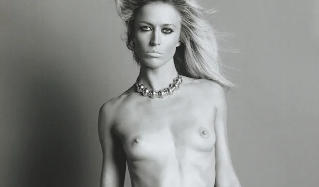 Black and white photo of a topless woman looking into the camera when standing in front of a plain background.