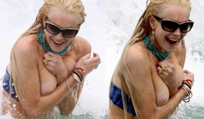 Lindsay Lohan with wet blonde hair and wearing sunglasses laughs while standing in water, holding her arms across her chest trying to keep her breasts contained within her blue bikini.