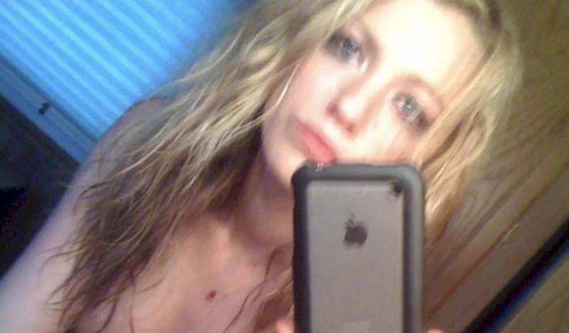 A woman with wet, wavy blonde hair takes a blurry selfie in a mirror, holding a black-cased iPhone. She has a neutral expression and is indoors, with a window and wooden wall visible in the background.