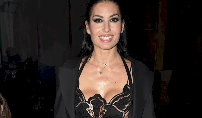 A woman with long dark hair smiles at the camera, wearing a black lace top and a black jacket draped over her shoulders, against a dark background.