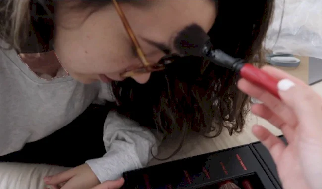 A girl wearing glasses leans forward as another hand applies makeup to their forehead with a brush. A makeup kit with red-handled brushes is visible in the foreground.