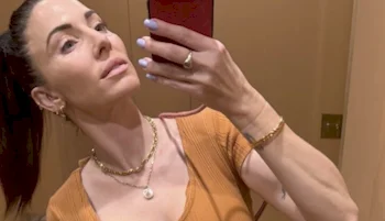 A woman with dark hair in a ponytail takes a mirror selfie indoors. She is wearing a ribbed orange top, gold jewelry, and has light blue nail polish. She holds her phone in one hand and looks slightly upward.