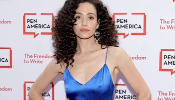 A woman standing in front of a white wall with repeated logos with hands on hips and wearing a blue satin dress.