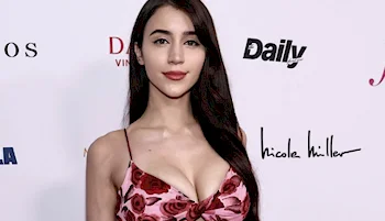 A woman with long dark hair wearing a pink floral dress poses in front of a step-and-repeat backdrop with logos, including Nicole Miller and Daily.