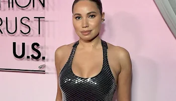 A woman with slicked-back hair wears a sleeveless, black sequin dress and poses in front of a pink backdrop with partially visible text.
