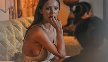 A woman in a sleeveless beige dress sits on a couch, looking concerned and biting her finger, while facing another person whose back is visible in the foreground. The setting appears to be a dimly lit room.