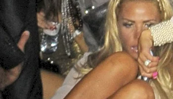 A woman with long blonde hair sits on the floor in a crowded environment, surrounded by people. She appears to be holding her legs and looking downward, with a pensive or indifferent expression.