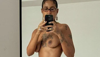 A woman taking a topless selfie. She is wearing glasses and is covered with tattoos.