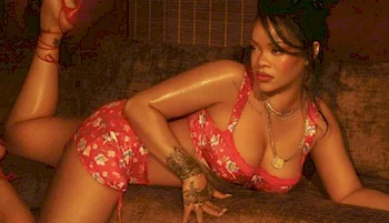 A woman with dark hair in an updo poses on a couch, wearing a matching floral pink bikini set. She has tattoos on her hand, gold jewelry, and is lit by a warm, soft light.