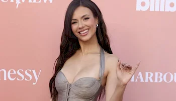 A woman with long dark hair in a strapless gray dress smiles and waves while posing in front of a pink backdrop with white text.