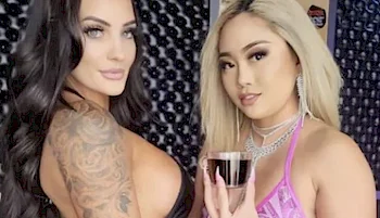 Two women posing together in front of a dark, patterned background. One has long dark hair and visible tattoos, the other has long blonde hair and wears a sparkly pink outfit, holding a small glass.