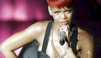 A woman with short red hair sings into a microphone on stage, wearing a black outfit with a gold chain necklace. The background is lit with pink and purple lighting.