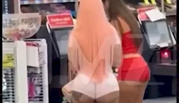 Two women are standing at a self-checkout counter in a store. One woman has long peach-colored hair and is wearing a light outfit, while the other has long brown hair and a red outfit. Their backs are facing the camera.