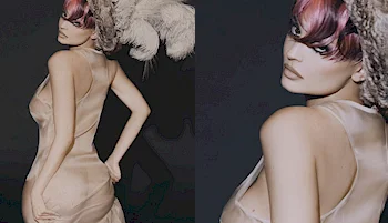 A person wearing a sheer, sleeveless dress and a dramatic feathered headpiece poses against a dark background. Their hair is styled with bangs, and they look over their shoulder with a serious expression.