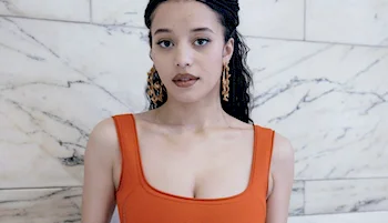 A woman with long, dark braided hair and statement earrings is wearing an orange sleeveless top. She stands in front of a white marble wall, looking directly at the camera with a neutral expression.