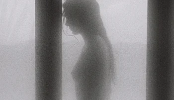 A blurred, grayscale silhouette of a topless woman with long hair stands between two vertical pillars, facing left. The background is hazy and indistinct, creating a mysterious, ethereal atmosphere.