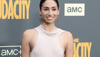 A woman wearing a sleeveless, light-colored dress smiles while standing in front of a gray backdrop with yellow and white text and AMC logos.