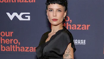 A woman with short dark hair and red lipstick poses in a black dress in front of a backdrop with orange and white text that reads our hero, balthazar and WG.
