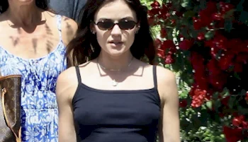 A woman wearing sunglasses and a black tank top walks outdoors, with another person in a blue patterned dress behind her and red flowers in the background.