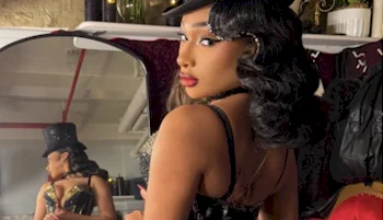 A woman with styled black hair and red lipstick wears a black outfit and hat, looking over her shoulder in front of a mirror reflecting her full costume in a vintage-themed room.