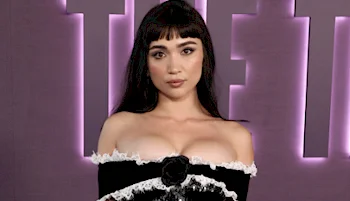 A woman with long dark hair and bangs poses in front of a purple backdrop with large, partially visible letters. She is wearing an off-shoulder black and white dress with lace trim.