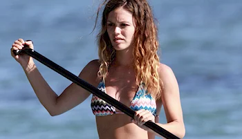 A woman in a patterned bikini holds a paddle while standing on water, with the ocean in the background.