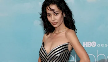 A woman in a silver and black striped strapless dress poses in front of a blue backdrop with the HBO logo partially visible.