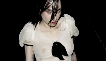 A pale young woman with dark hair covering her face, wearing a sheer white puffed-sleeve top with a dark bird motif, posed against a black background in a moody, dramatic style.