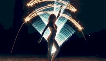 A nude woman stands outdoors at night, holding sparklers that create bright, curved light trails in the air. The background is dark, highlighting the glowing patterns around the figure.
