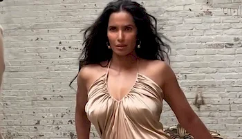 Padma Lakshmi wearing a shiny, draped beige dress stands confidently in front of a white brick wall.